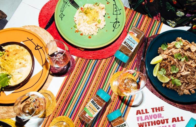 Cena mexicana repleta de platos, vino, mantel colorido, botellas de MEZCO y eslogan de sabor.